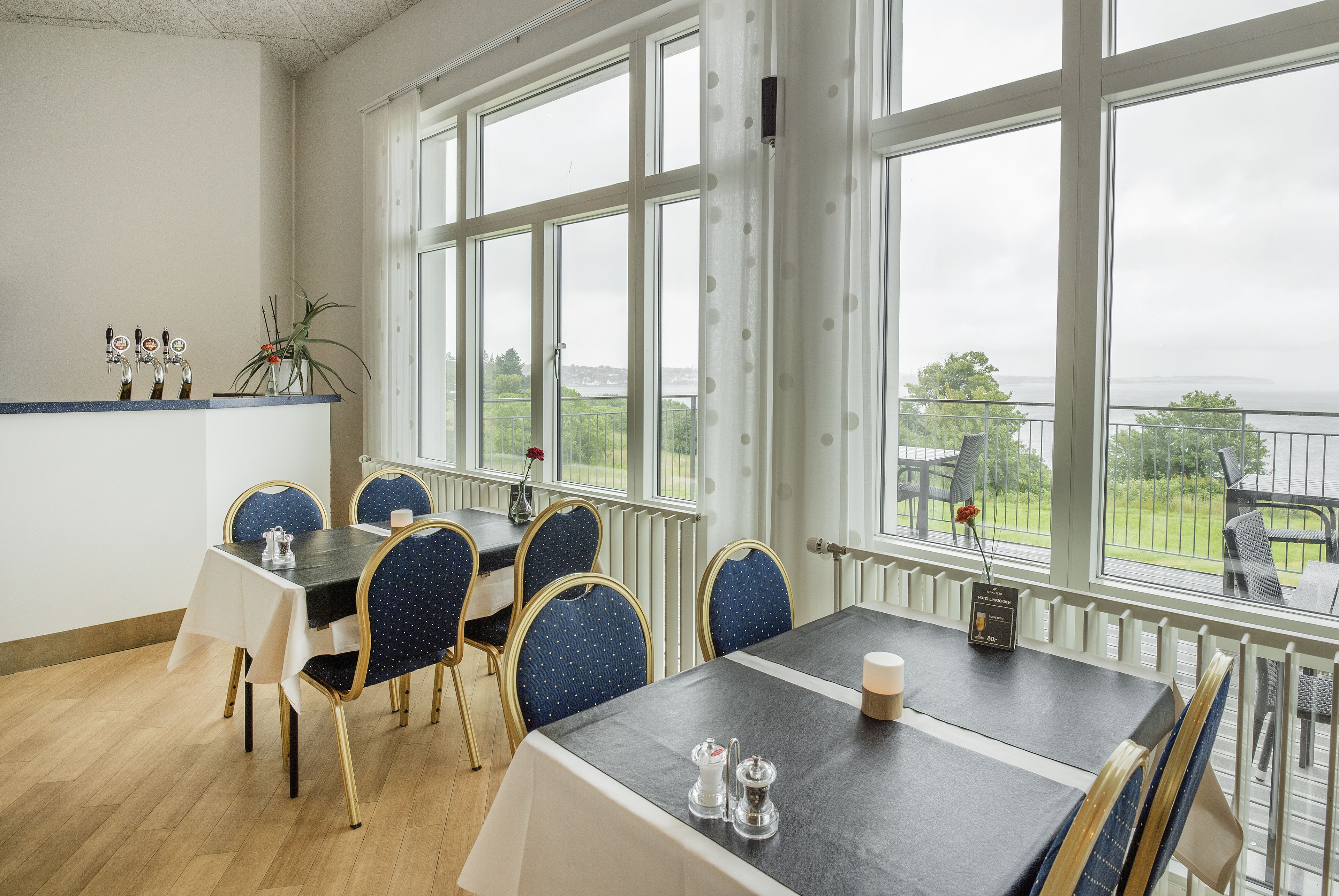 A dining area with a table and chairs, large windows with a view of greenery and a body of water, and a radiator under the windows.