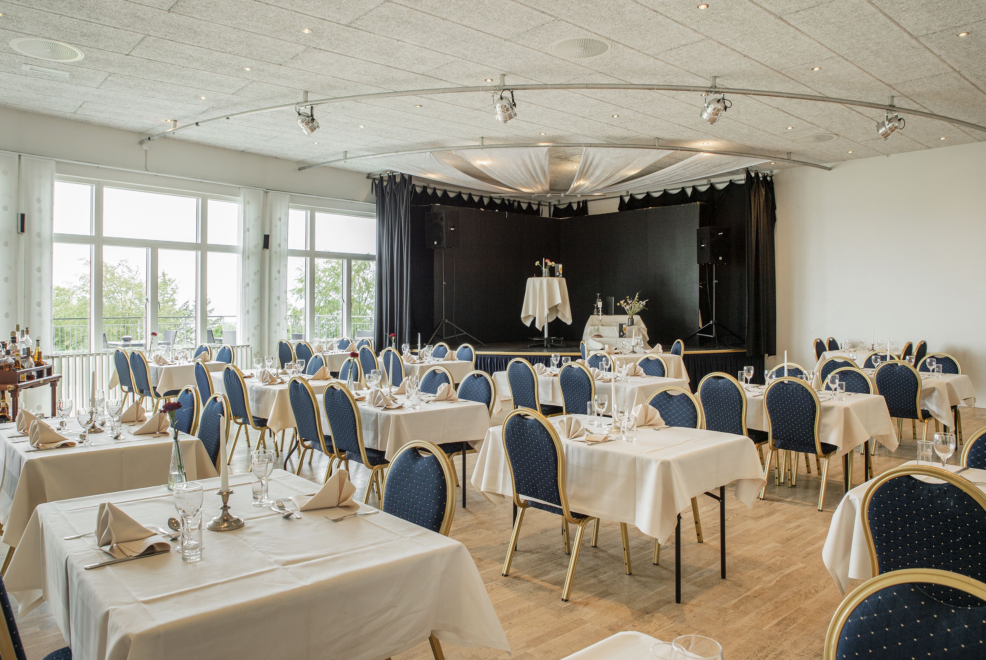 A banquet hall with multiple tables set for dining, a stage with curtains, and large windows allowing natural light.