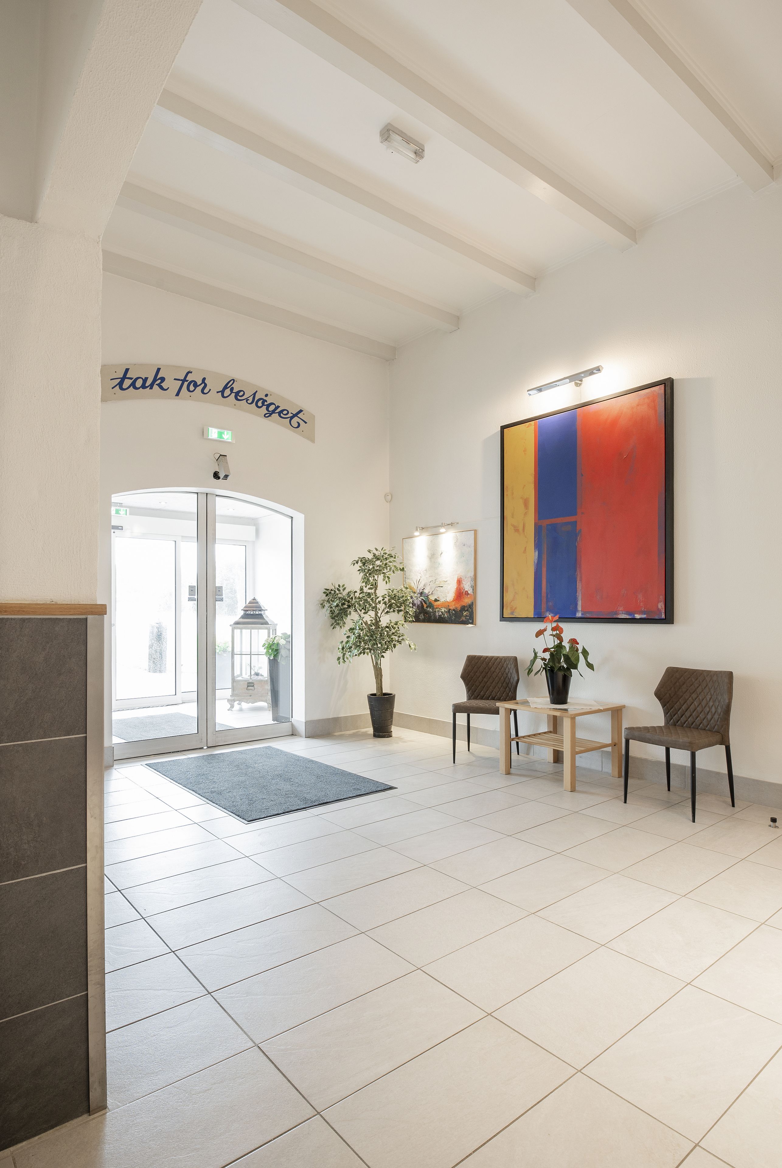 Een moderne lobby met een receptiebalie, twee stoelen, een potplant en een groot abstract schilderij.