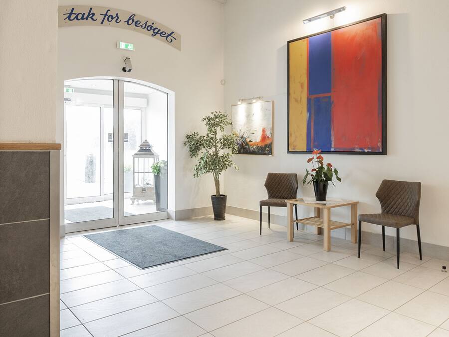 A modern lobby with a reception desk, two chairs, a potted plant, and a large abstract painting.