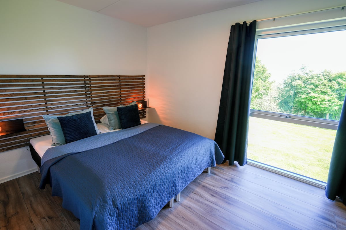 A bedroom with a wooden headboard, a large bed with a blue quilt, and a view of a grassy area through a window.