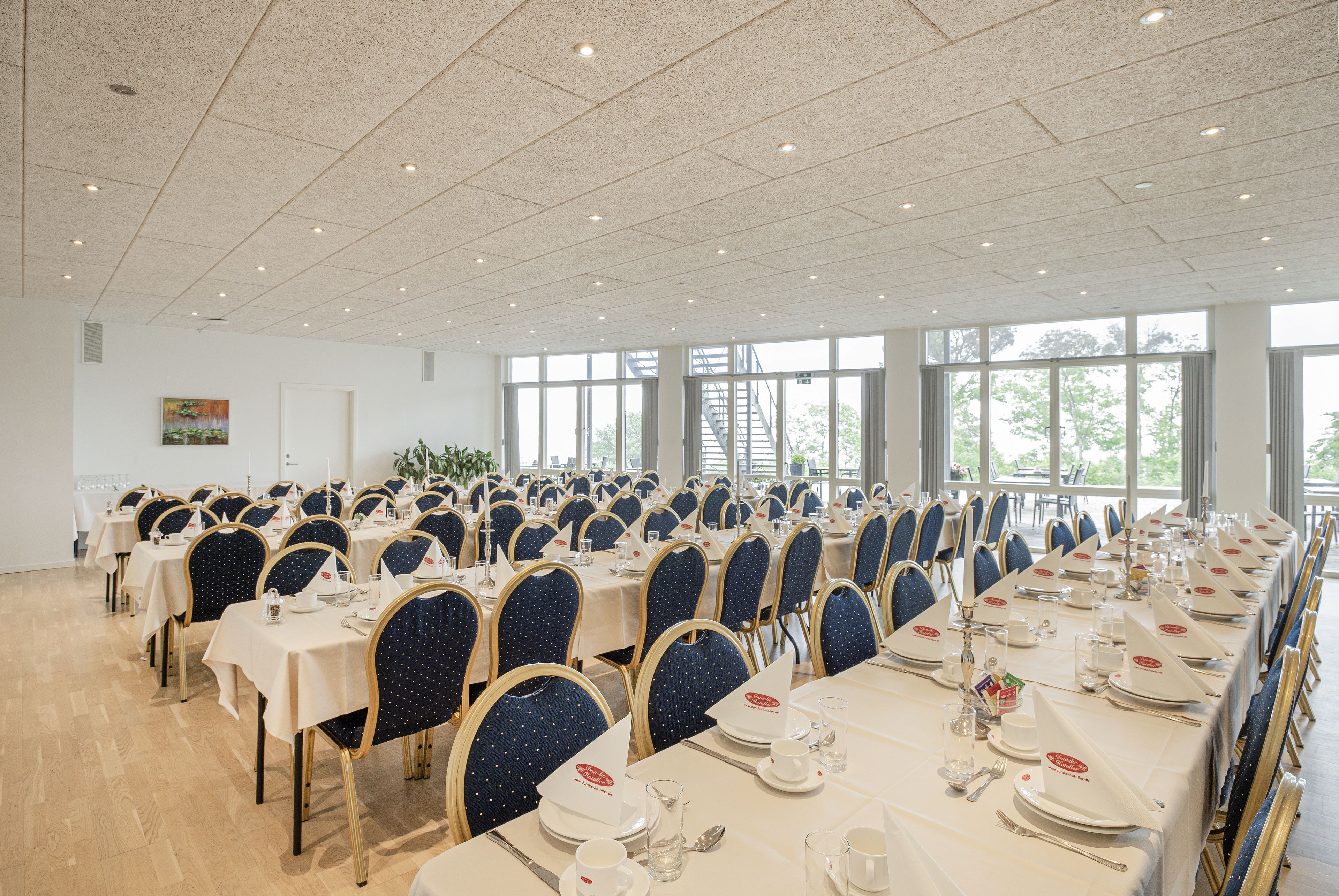 A banquet hall with rows of tables set for dining, chairs, and a staircase leading to an upper level.