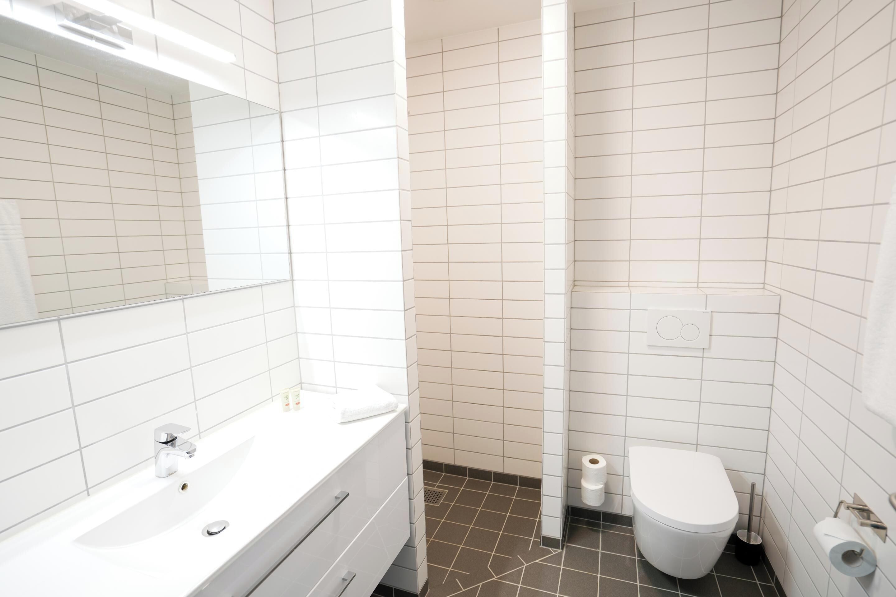 A modern bathroom with white tiles, a white sink with a chrome faucet, a toilet, and a shower area.