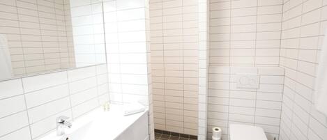 A modern bathroom with white tiles, a white sink with a chrome faucet, a toilet, and a shower area.