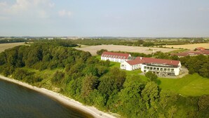 Un grand bâtiment blanc, surmonté d’un toit rouge, entouré d’une végétation luxuriante et bordé par un plan d’eau.