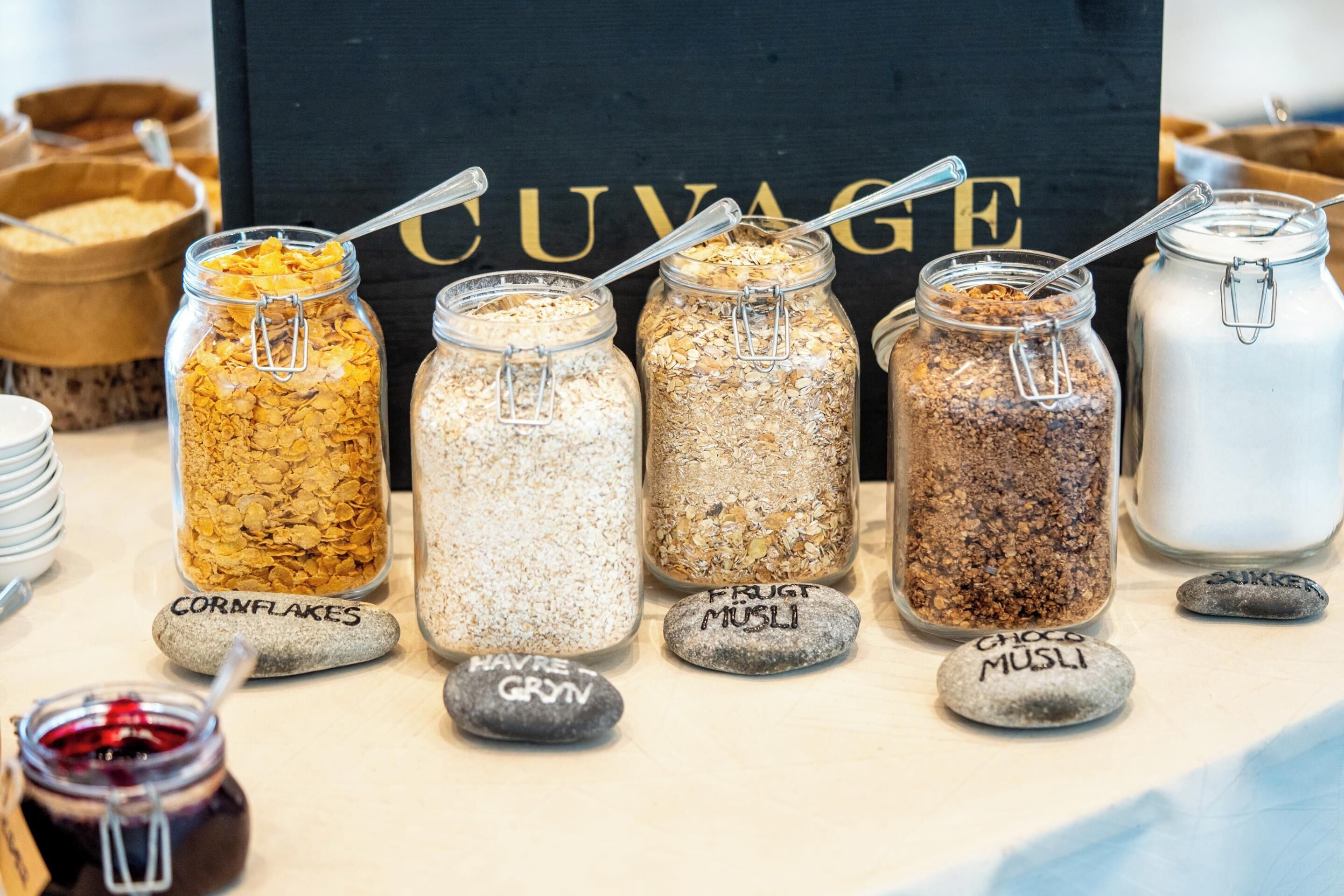 Diverse soorten levensmiddelen in potten, voorzien van etiketten met de inhoud, zoals cornflakes, fruitmuesli en chocolademuesli, tentoongesteld op een tafel.