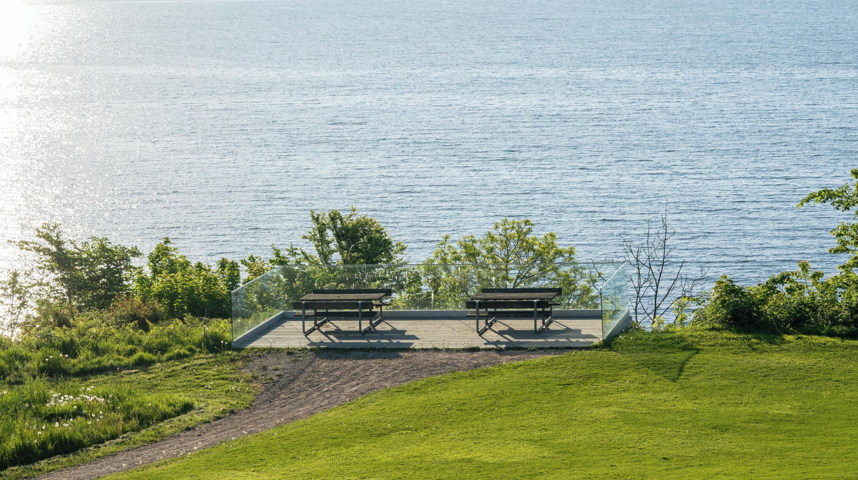 A lakeside deck with two wooden benches and a glass railing overlooking the water.
