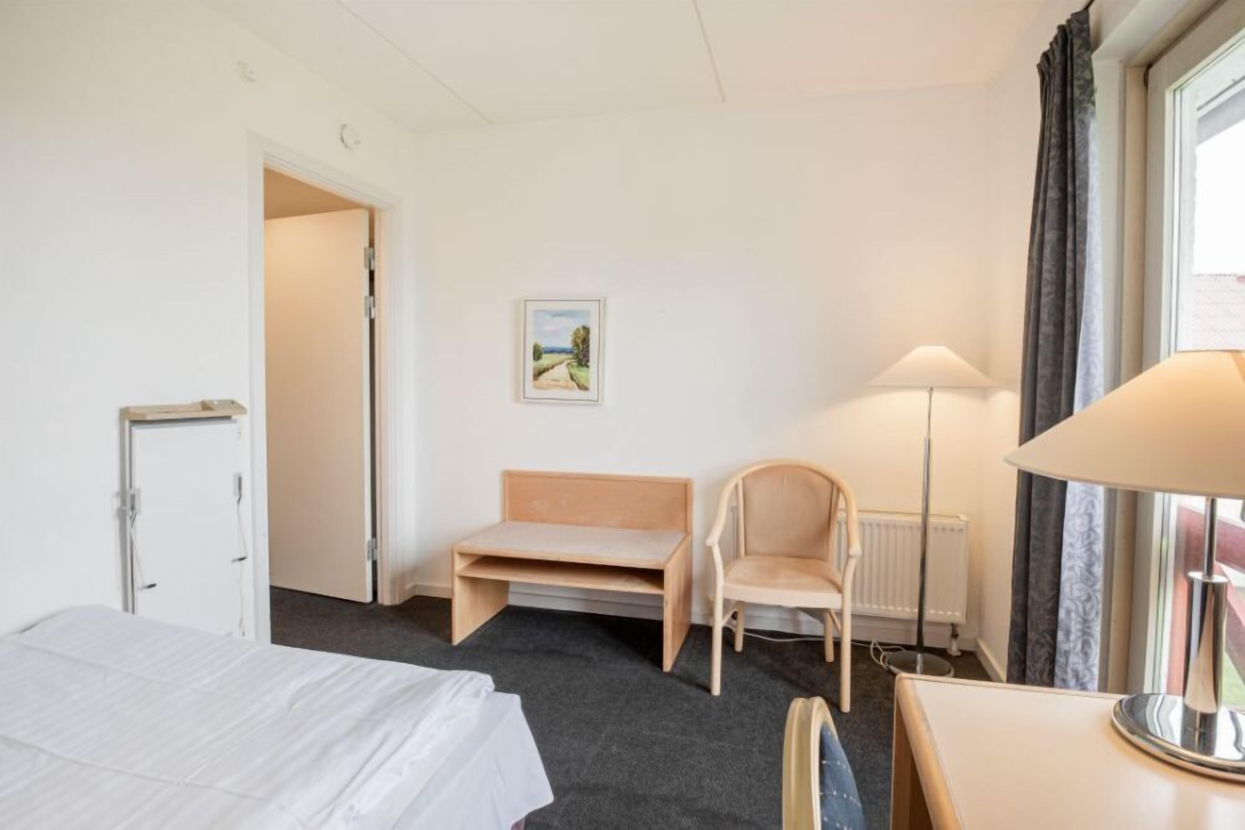 Une chambre d’hôtel avec un lit, un bureau, une chaise, une lampe, un radiateur et un tableau au mur.