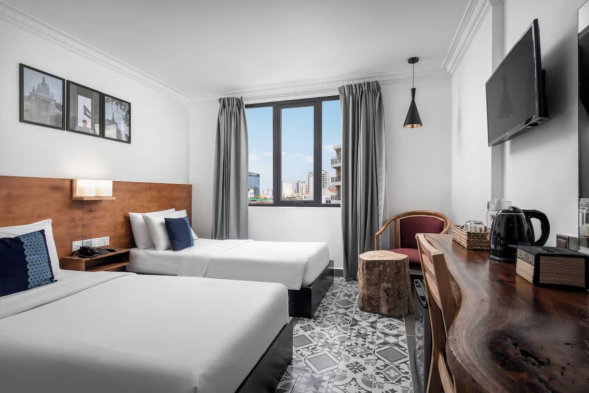 Une chambre d’hôtel avec deux lits, une tête de lit en bois, une grande fenêtre avec des rideaux, un téléviseur à écran plat, une table de chevet en bois avec une bouilloire et un tapis à motifs.
