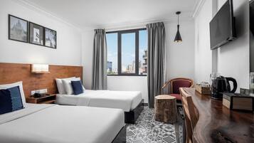 A hotel room with two beds, a wooden headboard, a large window with curtains, a flat-screen TV, a wooden side table with a kettle, and a patterned rug.