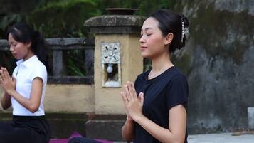 Two individuals seated on yoga mats outdoors, with a stone structure and greenery in the background.