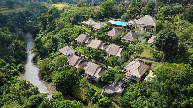 Luchtbeeld van een resort met meerdere gebouwen met rieten daken, een zwembad en een rivier die door het terrein stroomt.