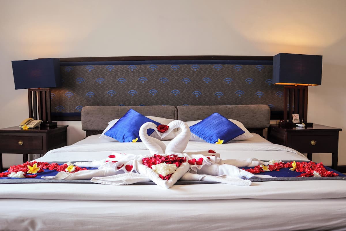A neatly made bed with white linens, a heart-shaped towel arrangement, and blue pillows.