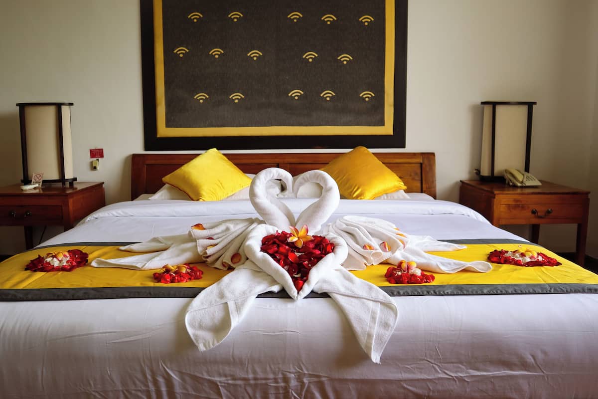 A hotel room with a bed featuring a heart-shaped towel arrangement and rose petals, yellow pillows, and a large framed picture on the wall.