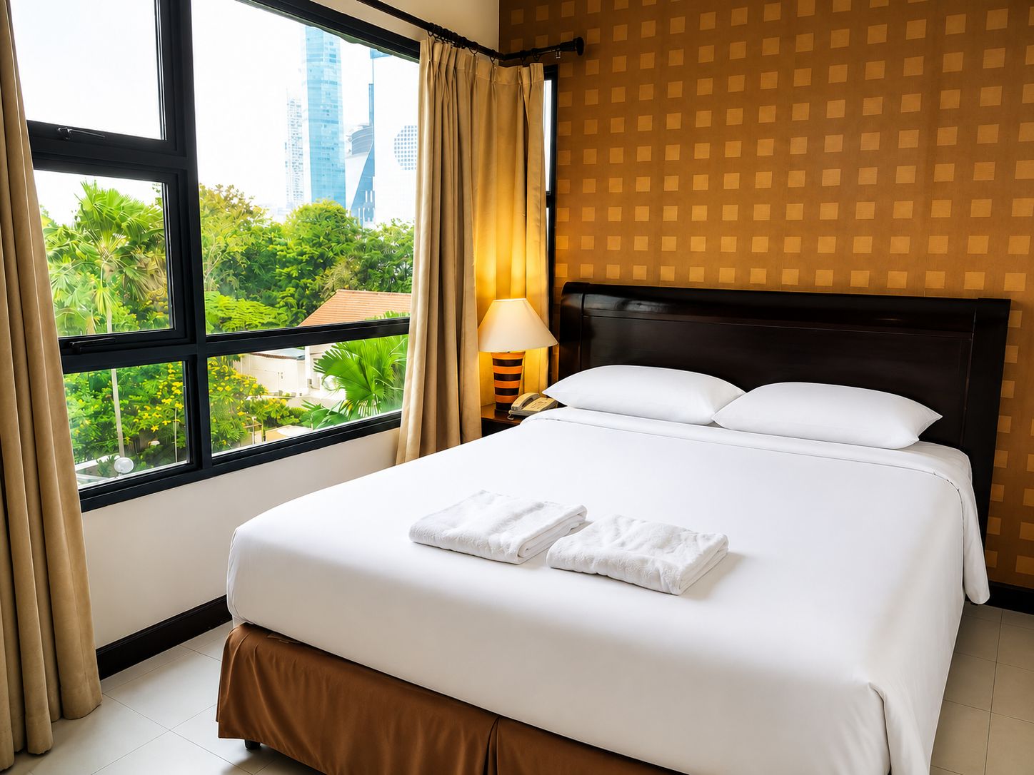 A hotel room with a large bed, a nightstand with a lamp, and a view of greenery and buildings through the window.