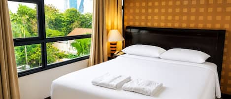 A hotel room with a large bed, a nightstand with a lamp, and a view of greenery and buildings through the window.