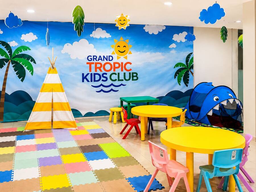Een kinderspeelruimte met een tent, tafels, stoelen en een muurschildering met de tekst 'GRAND TROPIC KIDS CLUB'.