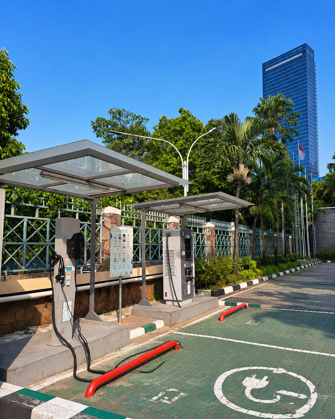 An electric vehicle charging station with a transparent roof, surrounded by greenery and a tall building in the background.