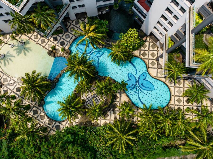 Vista aérea de un hotel con una piscina central rodeada de palmeras y un camino empedrado.