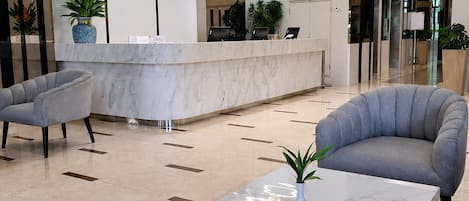 A modern lobby with a reception desk, two grey armchairs, a marble table, and a hanging light fixture.