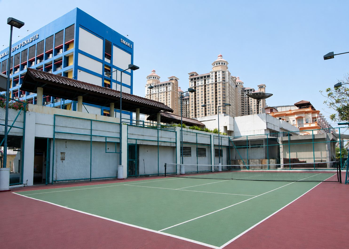 Una cancha de tenis con una red, rodeada de edificios, incluyendo una estructura azul con la señalización 'BPK PENABUR'.
