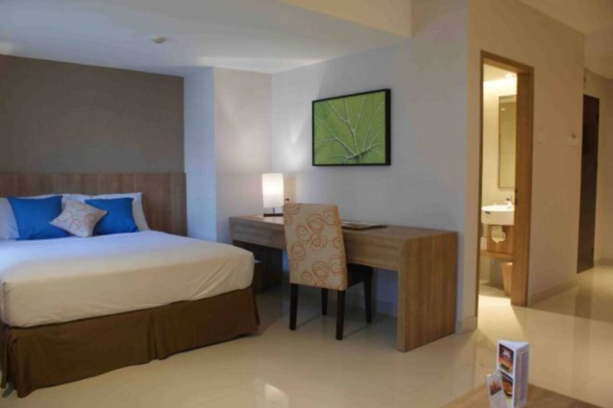 A hotel room with a bed, a desk, a chair, a lamp, and a framed picture on the wall.