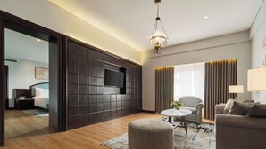 Een moderne hotelkamer met een houten vloer, een bank, een ronde salontafel, een televisie bevestigd aan een donkere paneelwand, en een groot raam met gordijnen.