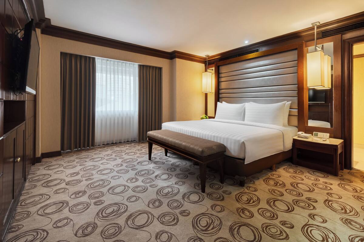 Une chambre d’hôtel avec un grand lit, une banquette, une table de chevet avec une lampe, une télévision et une grande fenêtre avec des rideaux.