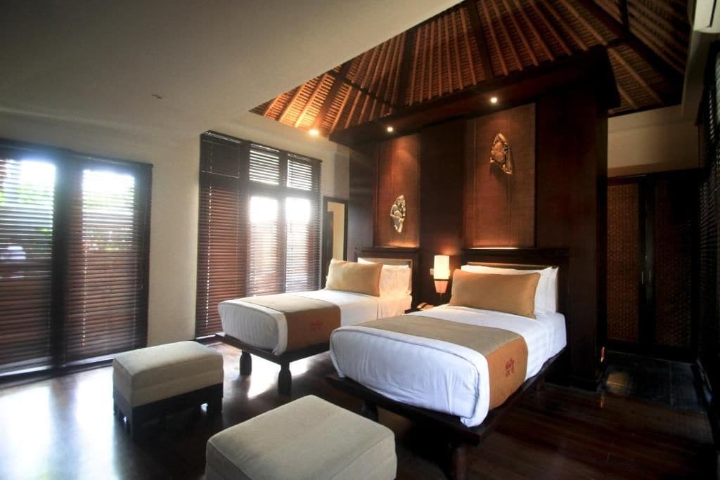 A hotel room with two beds, a large window with blinds, a wooden ceiling, and a small table.