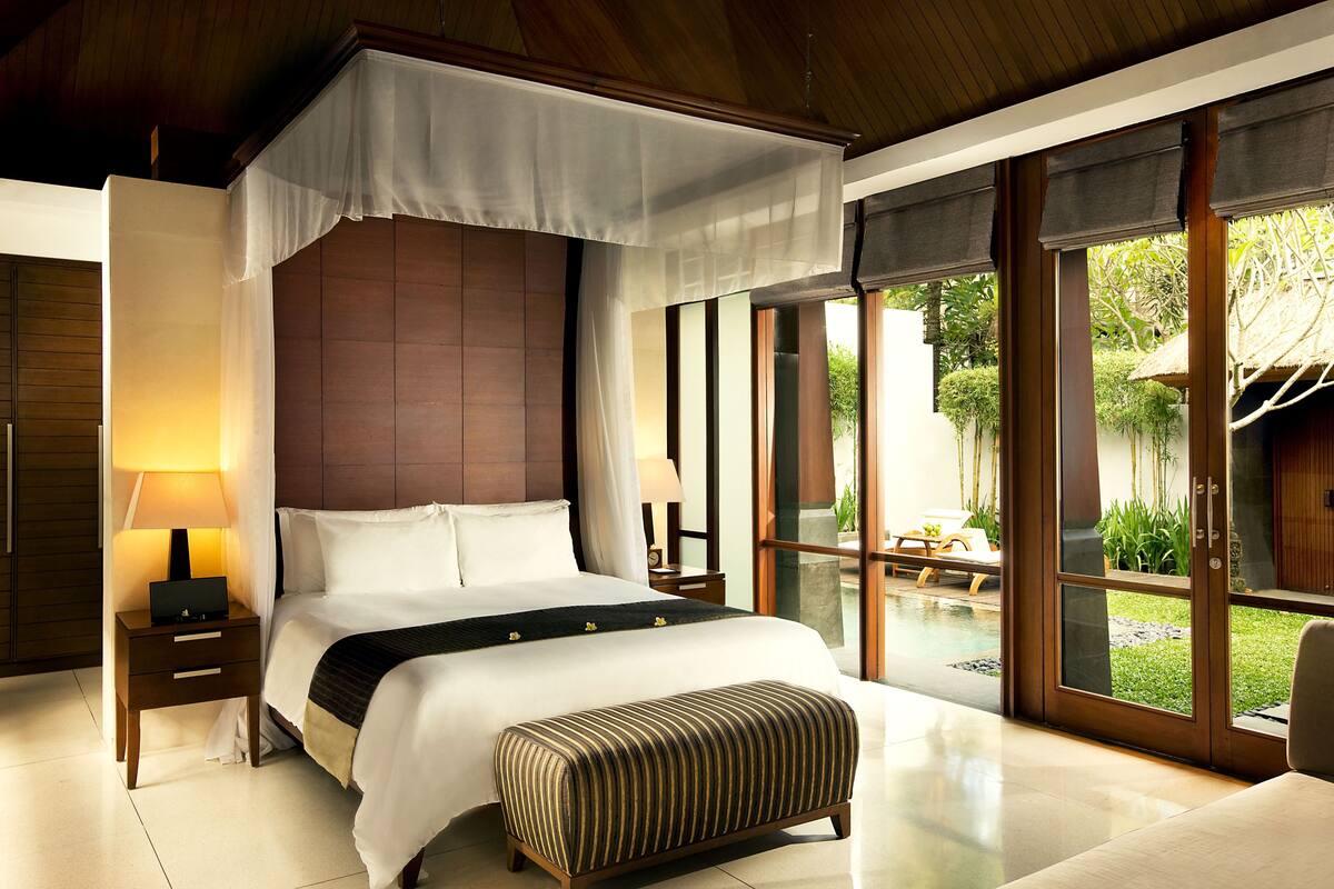 A spacious bedroom with a large bed, a bedside table with a lamp, a bench, and a view of a pool and garden through large windows.