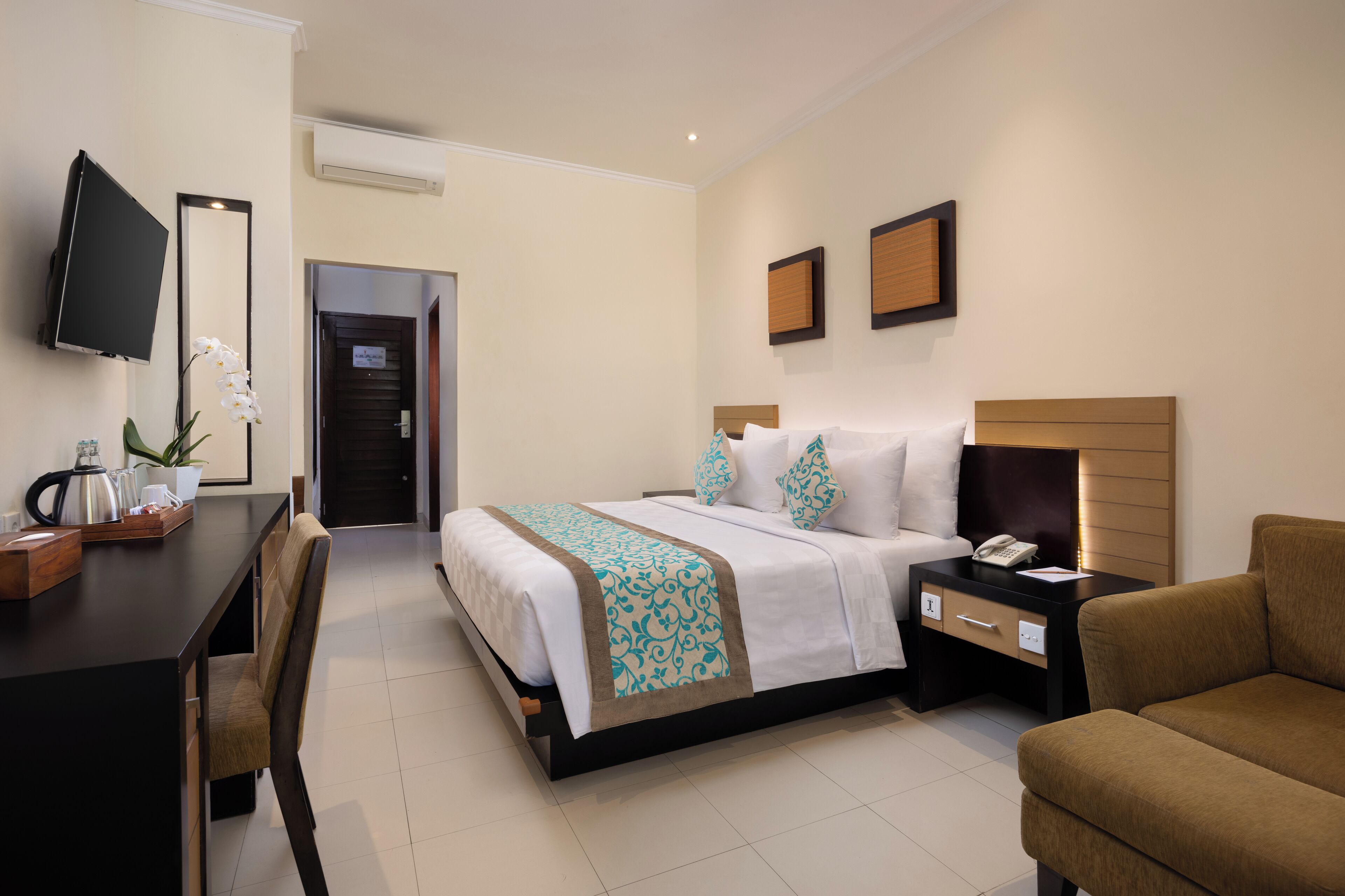 A hotel room with a bed, bedside table, chair, desk, TV, and a potted plant.