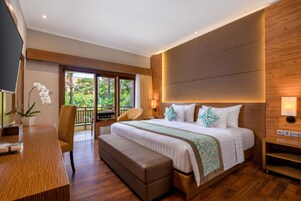 A hotel room with a large bed, a desk, a bench, a TV, and a balcony with a view of greenery.