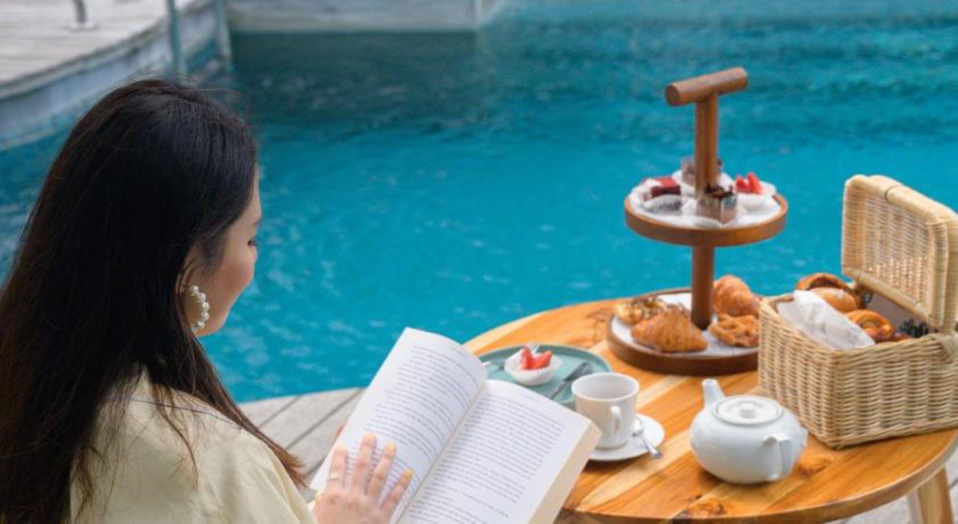 Una persona leyendo un libro junto a una mesa en la piscina, con un juego de té, pasteles y una bandeja de frutas.
