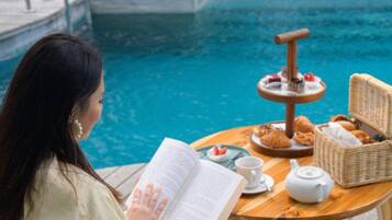 Una persona leyendo un libro junto a una mesa en la piscina, con un juego de té, pasteles y una bandeja de frutas.