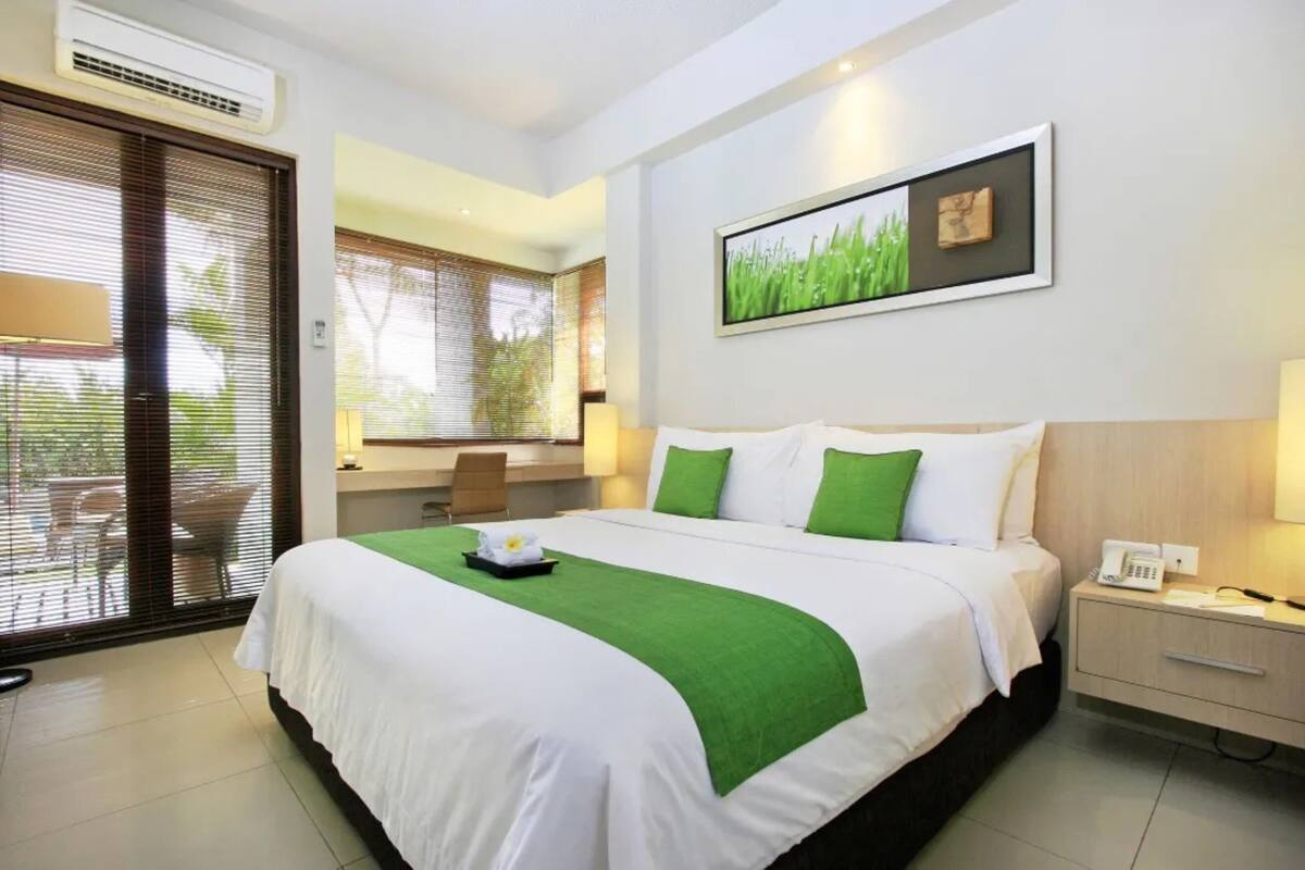 A hotel room with a bed, a desk, and a chair. There is a window with blinds, a picture on the wall, and a phone on the nightstand.