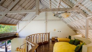 A spacious room with a high ceiling, wooden beams, a yellow sofa with green cushions, a wooden staircase, and a view of the outdoors.