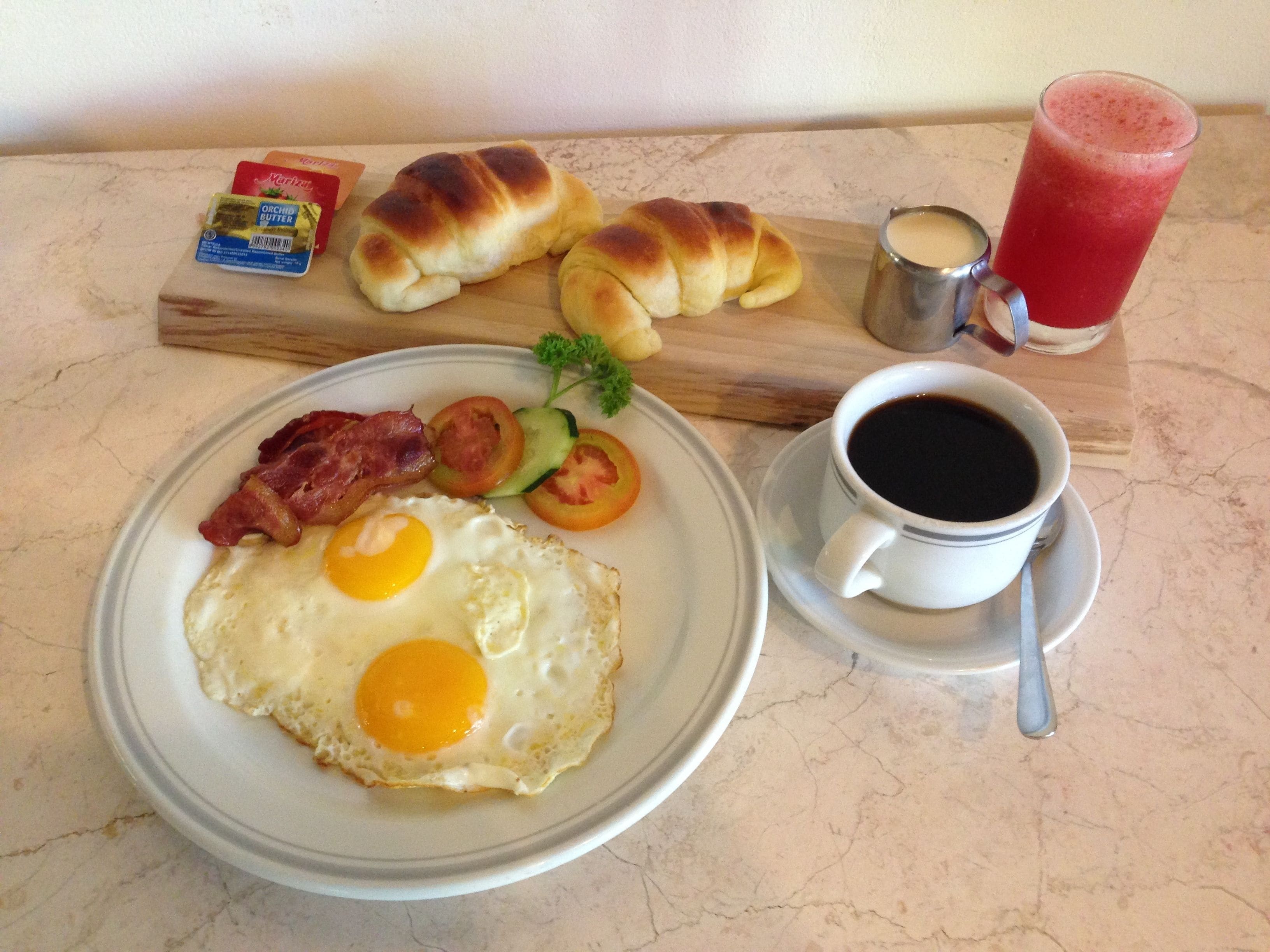 A breakfast plate with eggs, bacon, tomatoes, and a croissant, along with a cup of coffee and a glass of juice on a marble table.