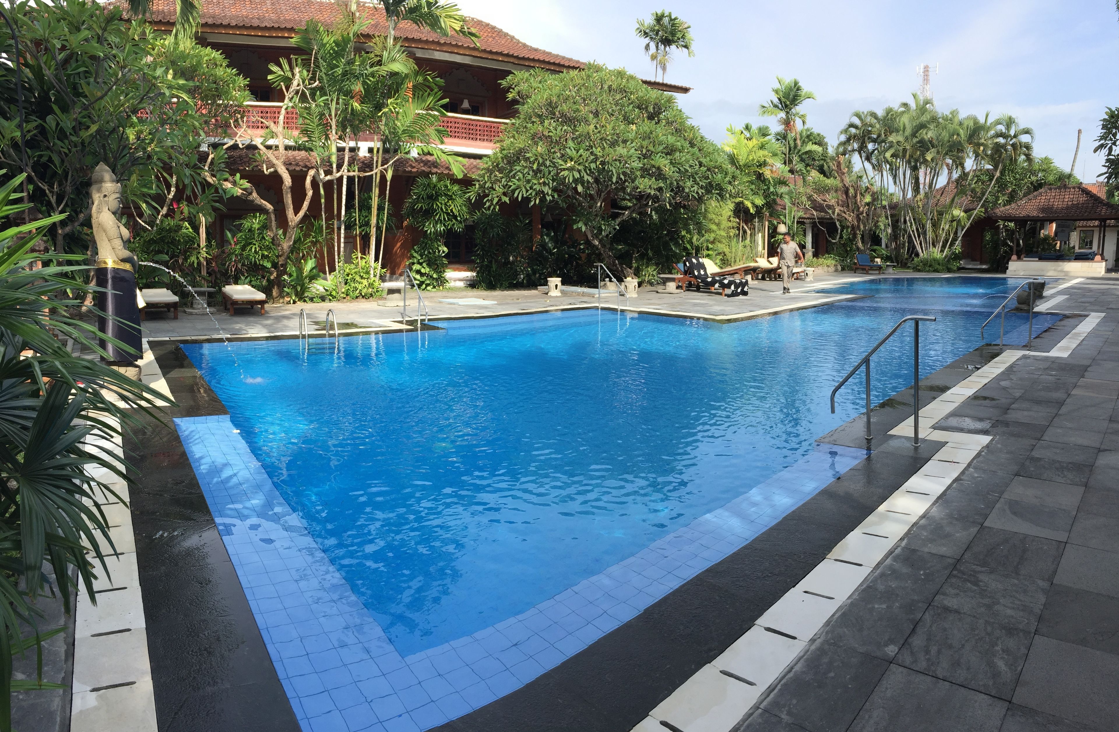 En swimmingpool med flisekant, omgivet af frodig vegetation og en bygning med flisebelagt tag.