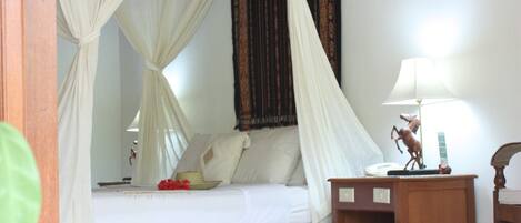 A room with a bed under a white canopy, a bedside table with a lamp and a decorative item, and a wooden chair.