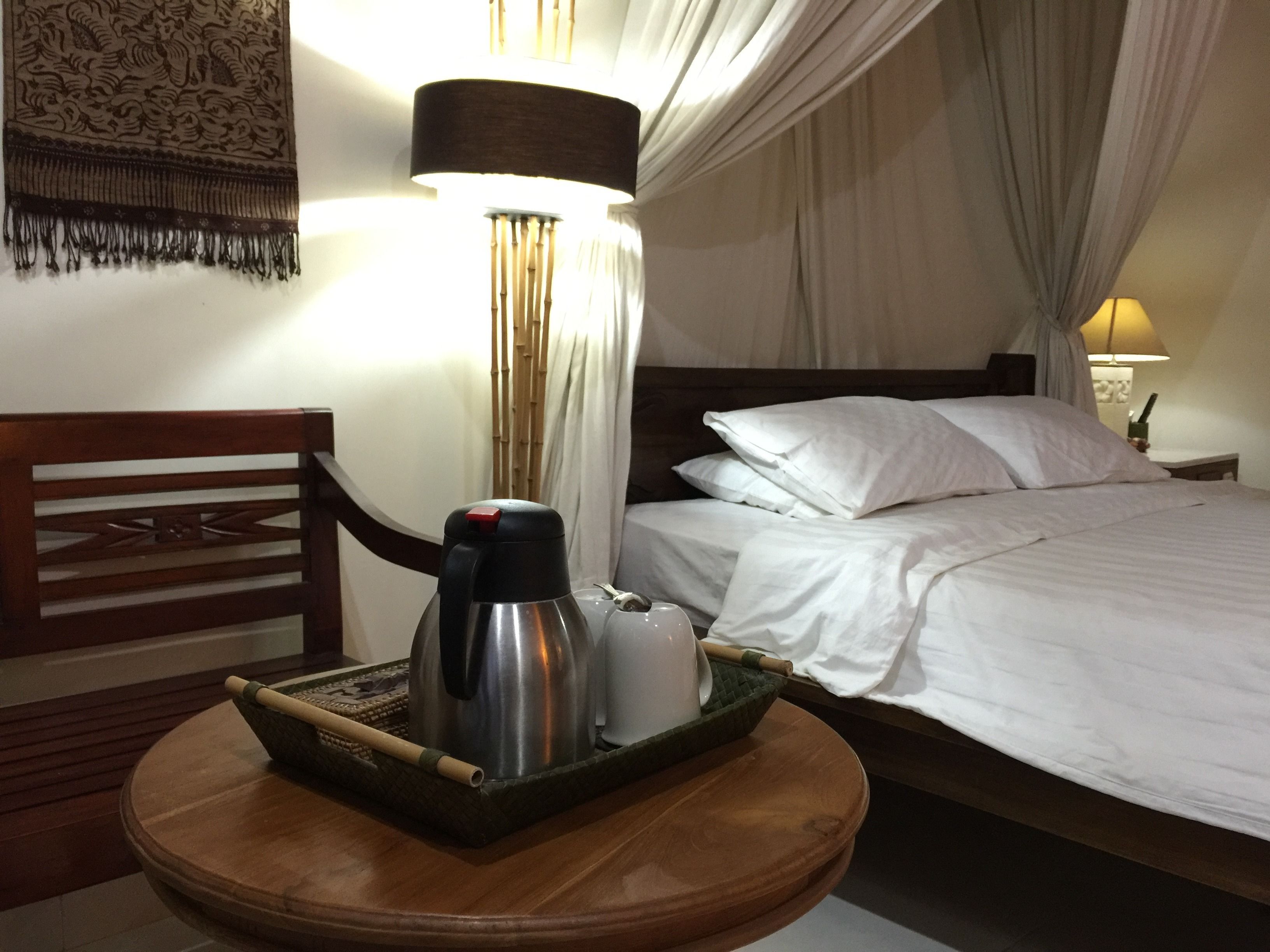 A hotel room with a bed, a bedside table with a tray containing a kettle, a cup, and a bamboo stick.