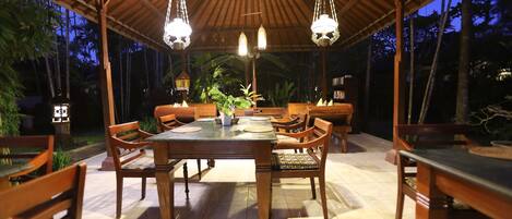 An outdoor dining area with a wooden table and chairs, surrounded by a covered roof structure with hanging lights.