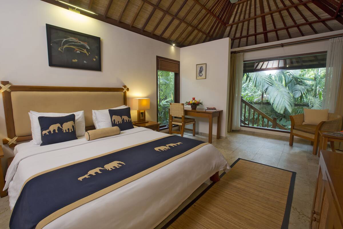 A hotel room with a bed, a desk, a chair, and a balcony with a view of greenery.