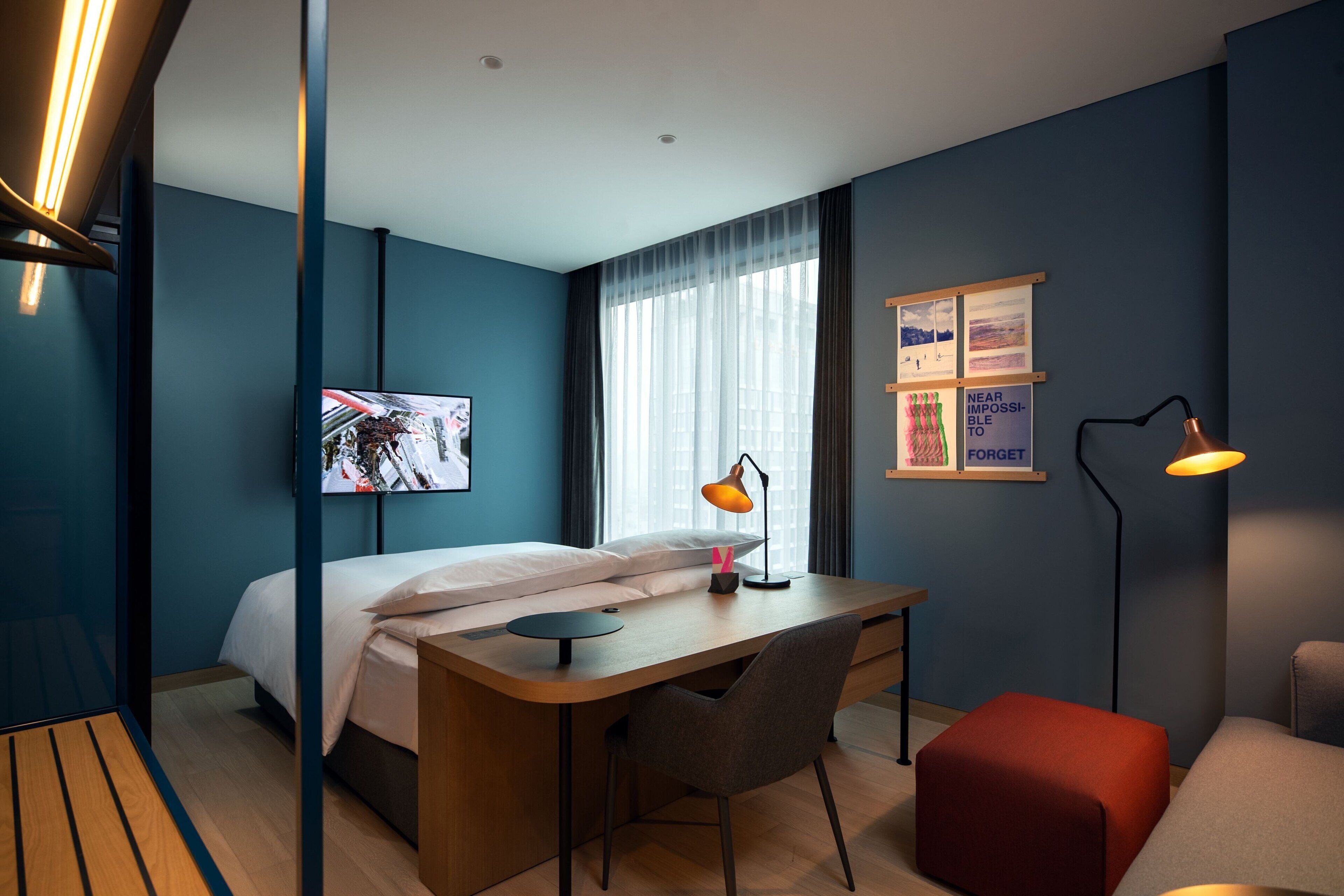 Una habitación de hotel moderna con cama, escritorio, silla y un otomán rojo.