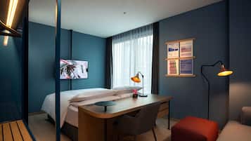 A modern hotel room with a bed, desk, chair, and a red ottoman.