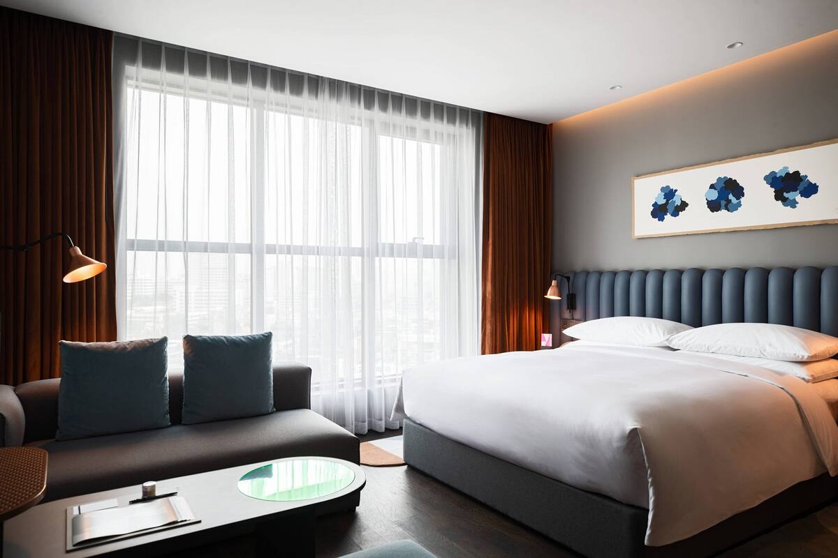 A modern hotel room with a large bed, a sofa, a small table, and a large window with sheer curtains.