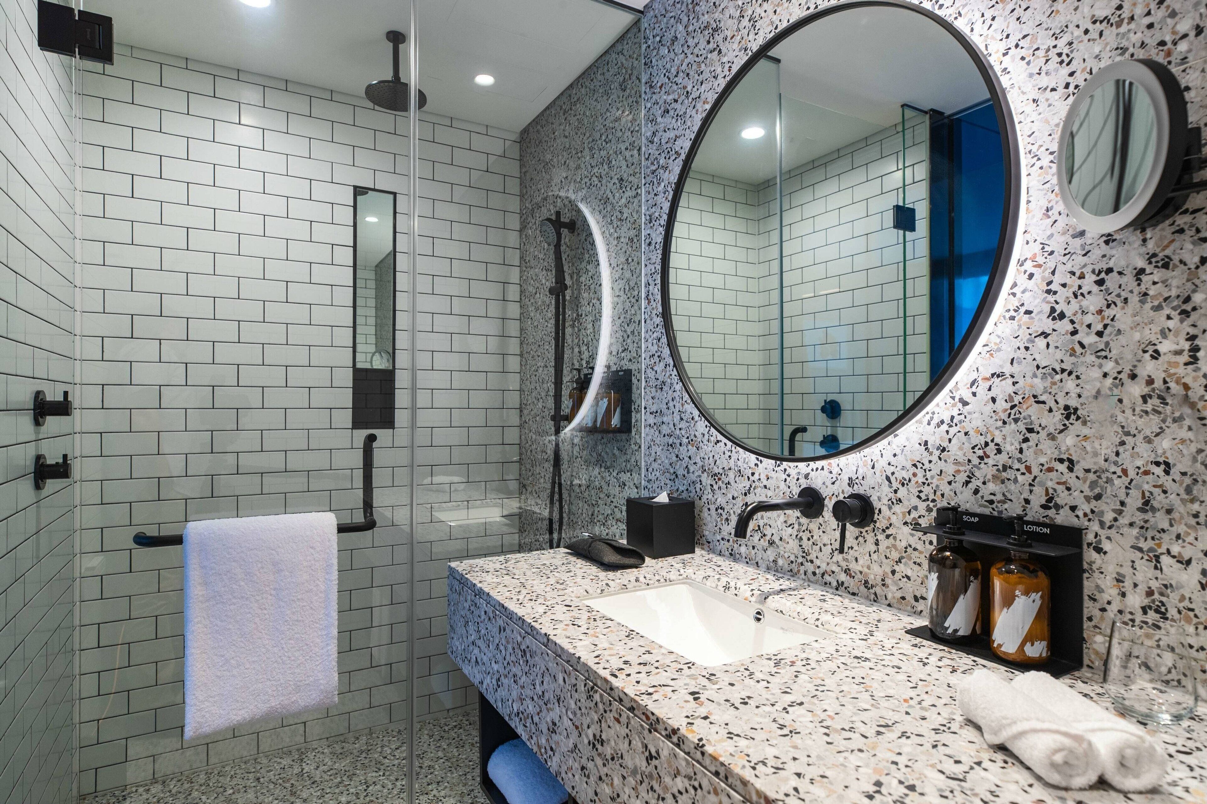 A modern bathroom with white and speckled tiles, a large round mirror, a rectangular sink, and two soap dispensers.