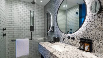 A modern bathroom with white and speckled tiles, a large round mirror, a rectangular sink, and two soap dispensers.