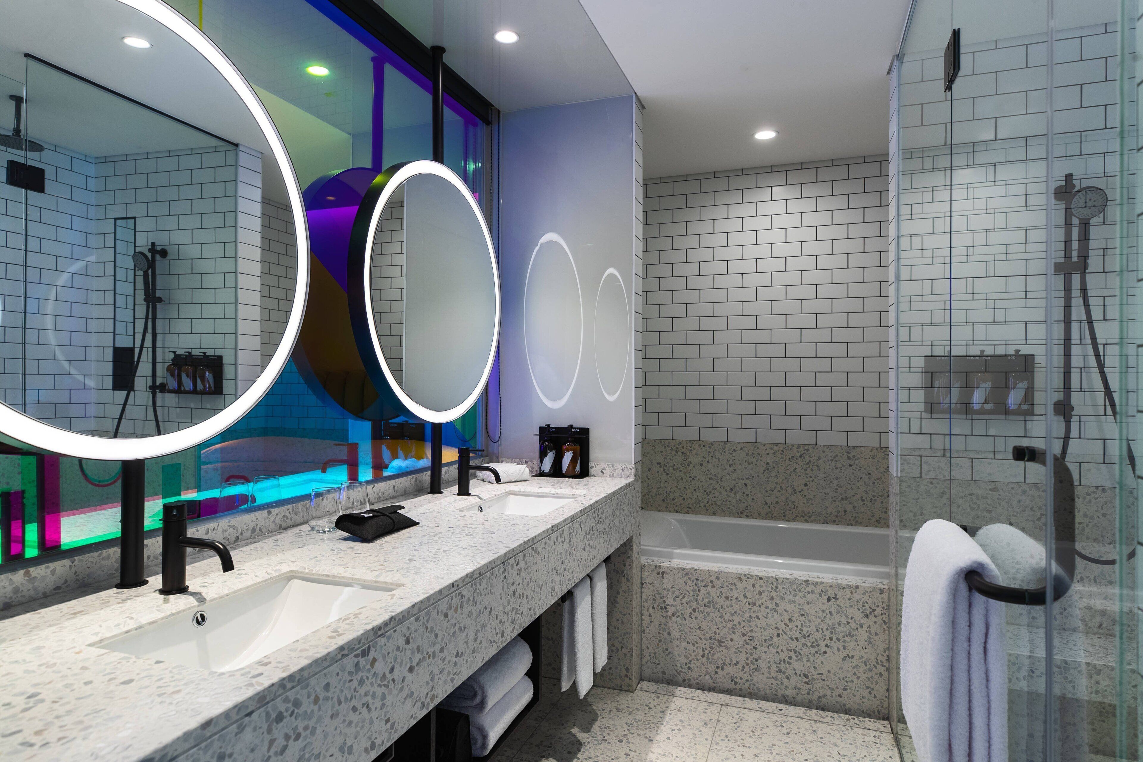Un baño moderno con dos lavamanos, un espejo grande con luces circulares, cabina de ducha de vidrio y un reloj visible en la pared.