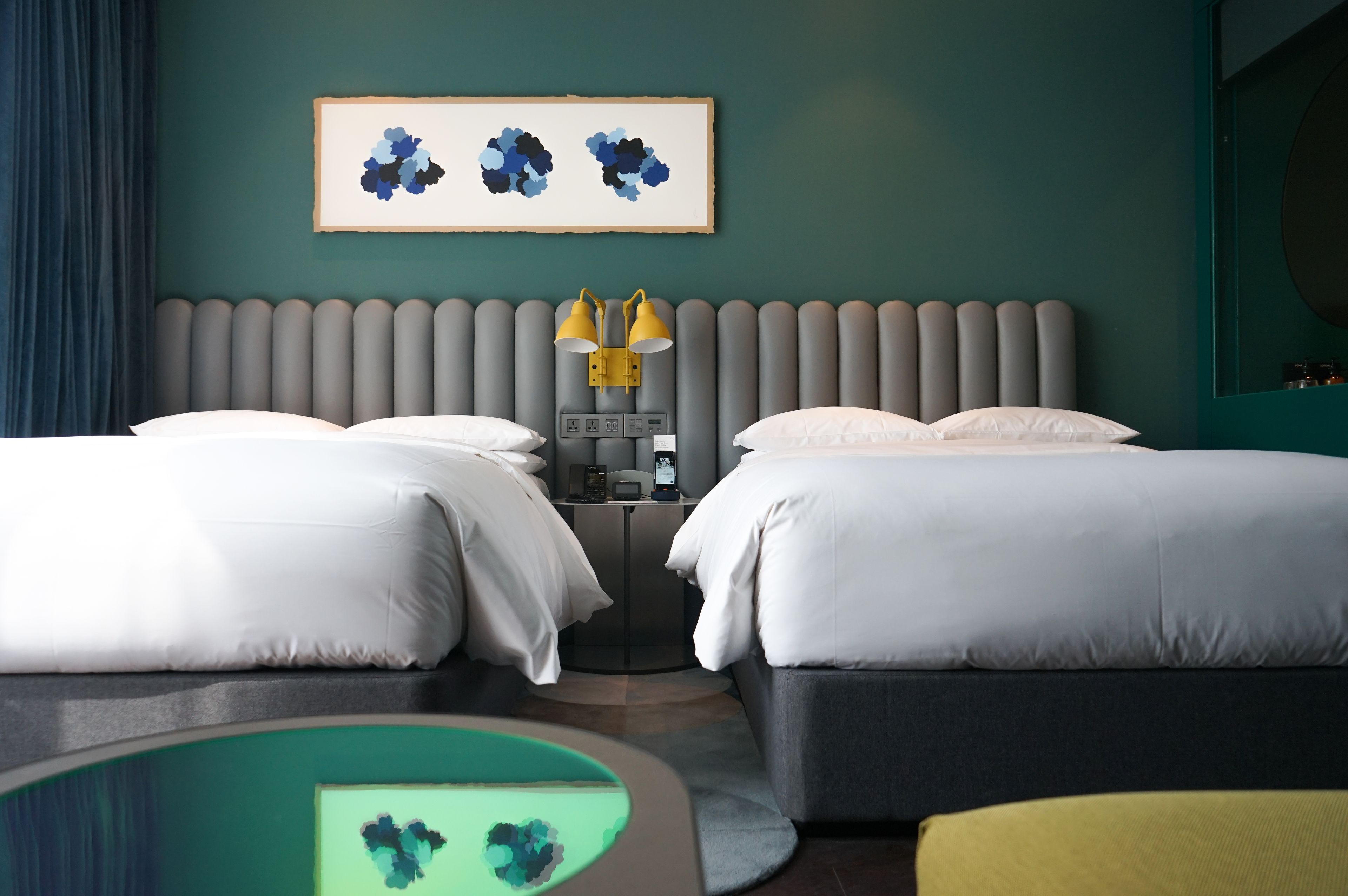 A hotel room with two beds, a bedside table with lamps, a framed artwork on the wall, and a round table with a green top.