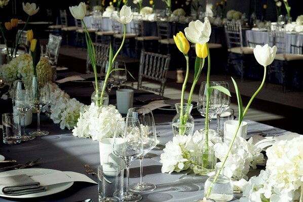 A banquet hall with tables set for dining, featuring white and yellow tulips and white flowers in glass vases.